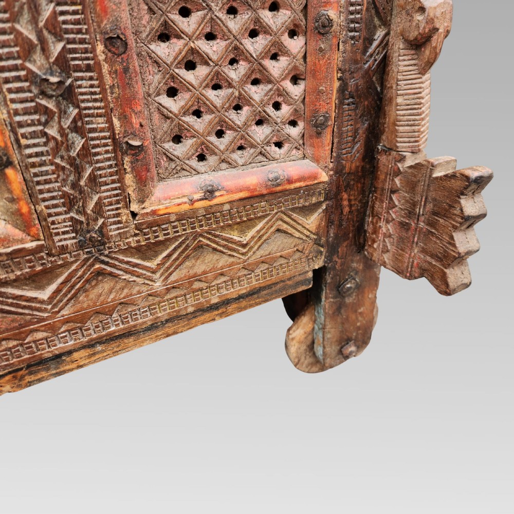 An intricately carved antique Indian teak Damchiya dowry chest featuring painted glass panels, horse head bracket carvings, metal studs, and small wooden wheels. View XIII