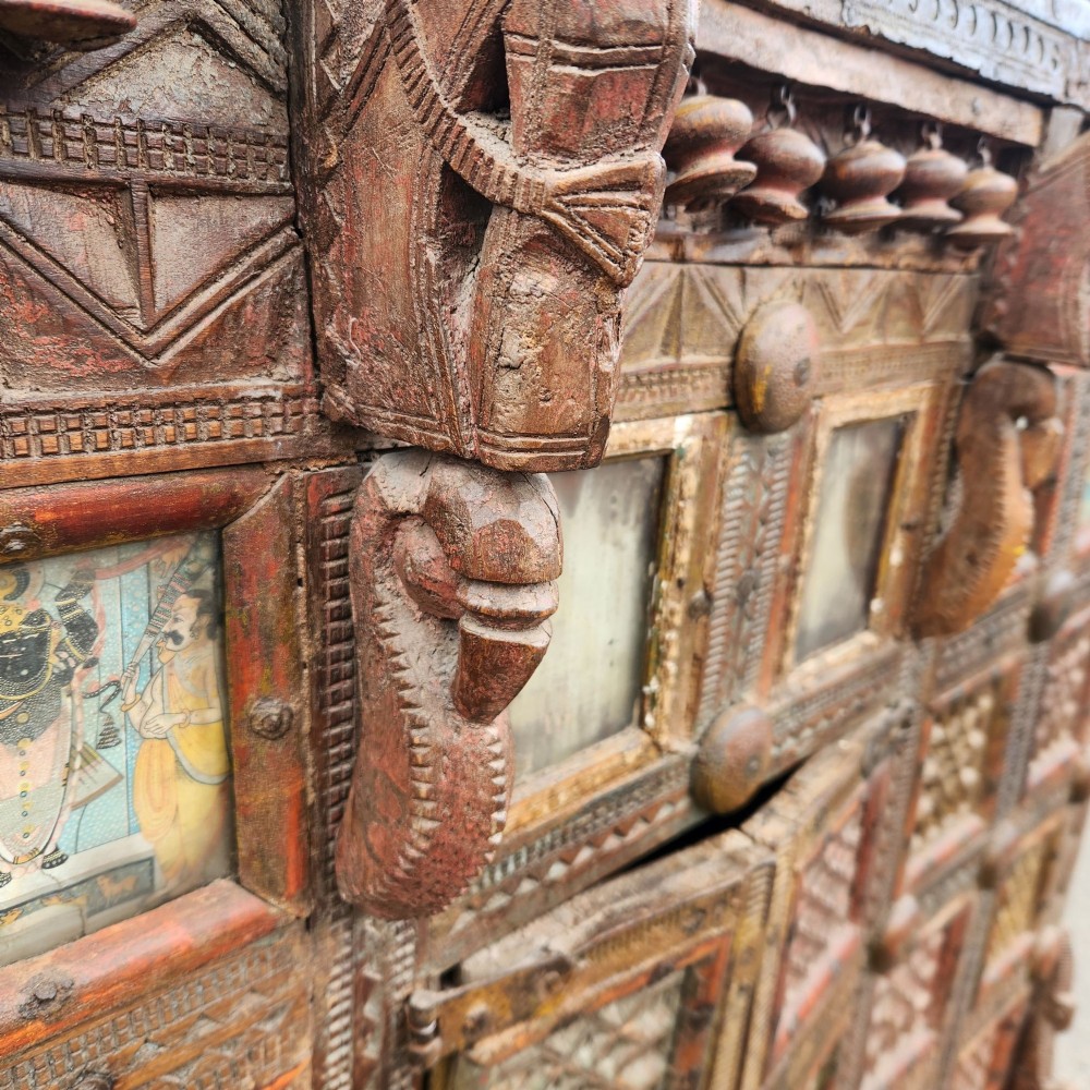 An intricately carved antique Indian teak Damchiya dowry chest featuring painted glass panels, horse head bracket carvings, metal studs, and small wooden wheels. View XIV