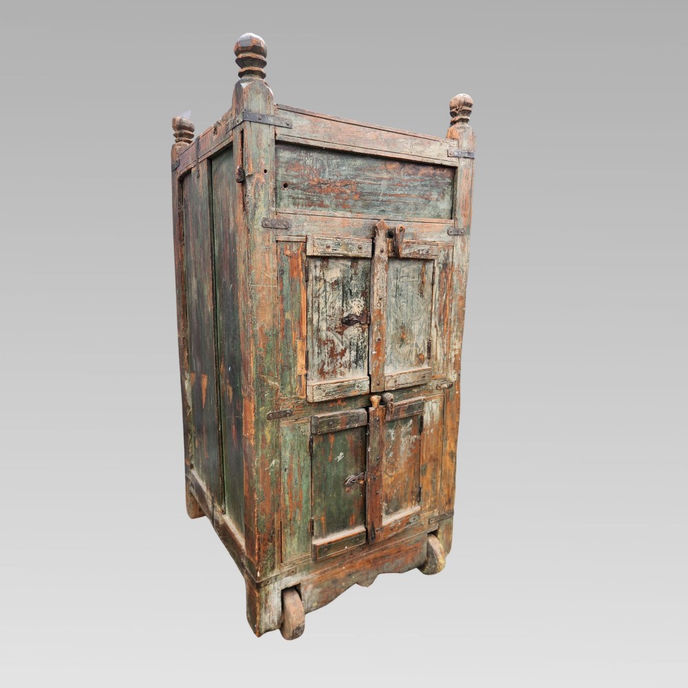 A rustic wooden Indian almirah storage cupboard with distressed green paint, double panel doors, iron fittings, turned corner finials, and wooden wheels.