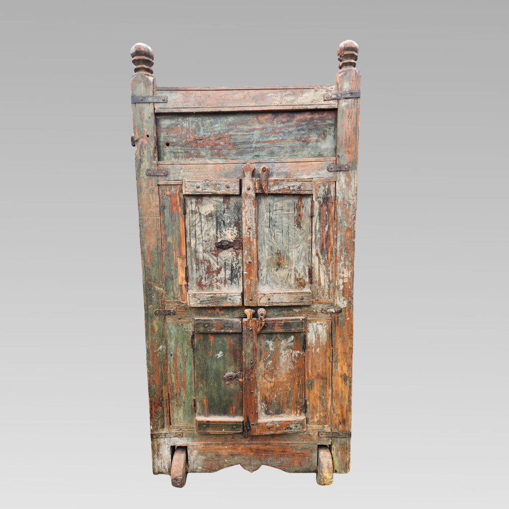 A rustic wooden Indian almirah storage cupboard with distressed green paint, double panel doors, iron fittings, turned corner finials, and wooden wheels. View II