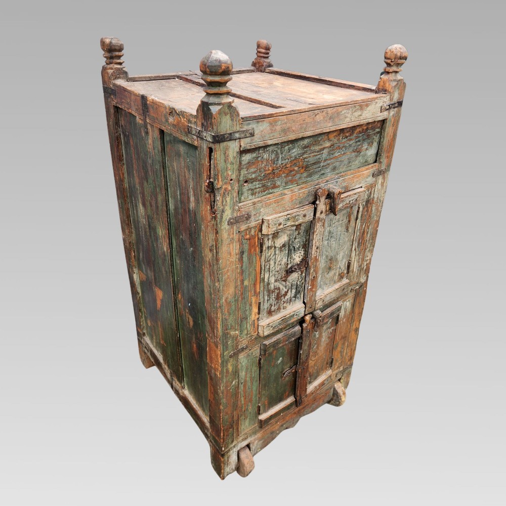 A rustic wooden Indian almirah storage cupboard with distressed green paint, double panel doors, iron fittings, turned corner finials, and wooden wheels. View III