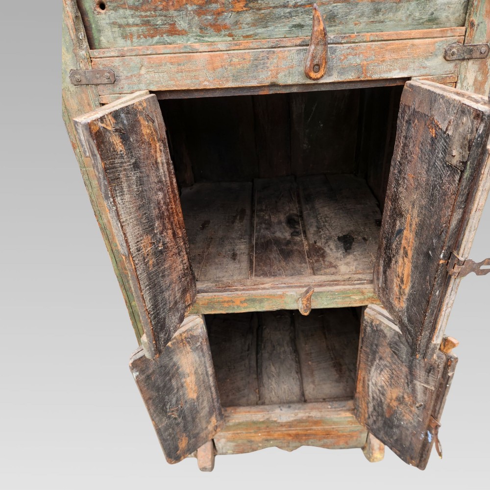 A rustic wooden Indian almirah storage cupboard with distressed green paint, double panel doors, iron fittings, turned corner finials, and wooden wheels. View IV
