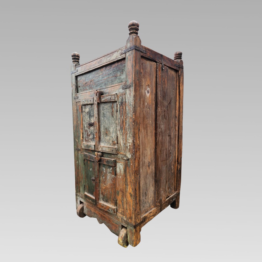 A rustic wooden Indian almirah storage cupboard with distressed green paint, double panel doors, iron fittings, turned corner finials, and wooden wheels. View V