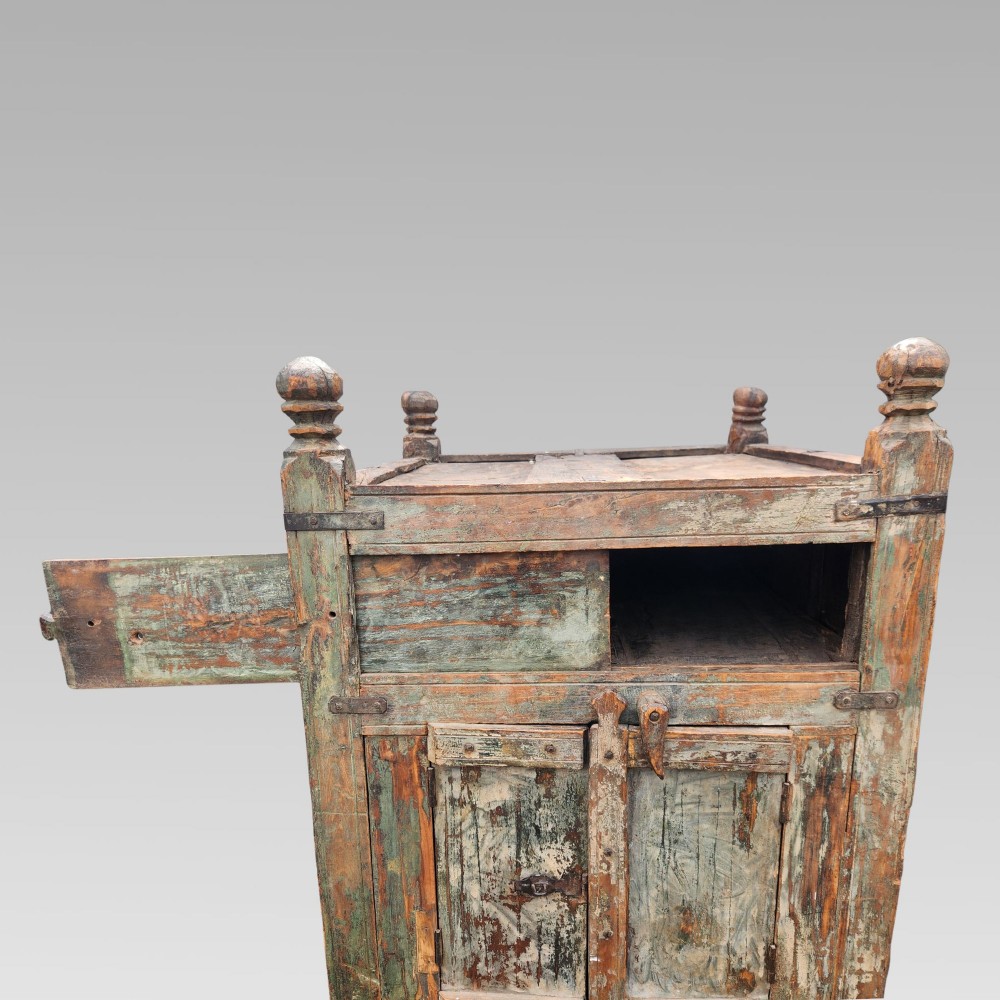 A rustic wooden Indian almirah storage cupboard with distressed green paint, double panel doors, iron fittings, turned corner finials, and wooden wheels. View VI