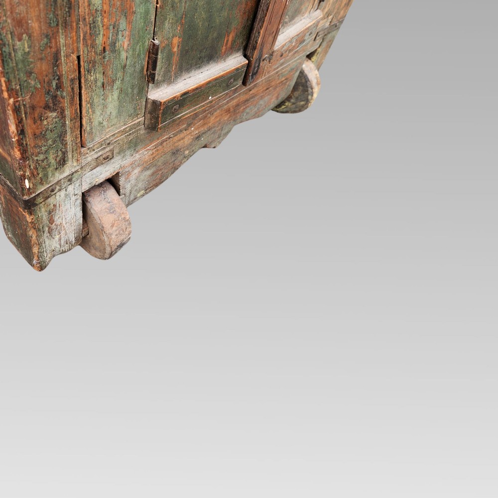 A rustic wooden Indian almirah storage cupboard with distressed green paint, double panel doors, iron fittings, turned corner finials, and wooden wheels. View VII