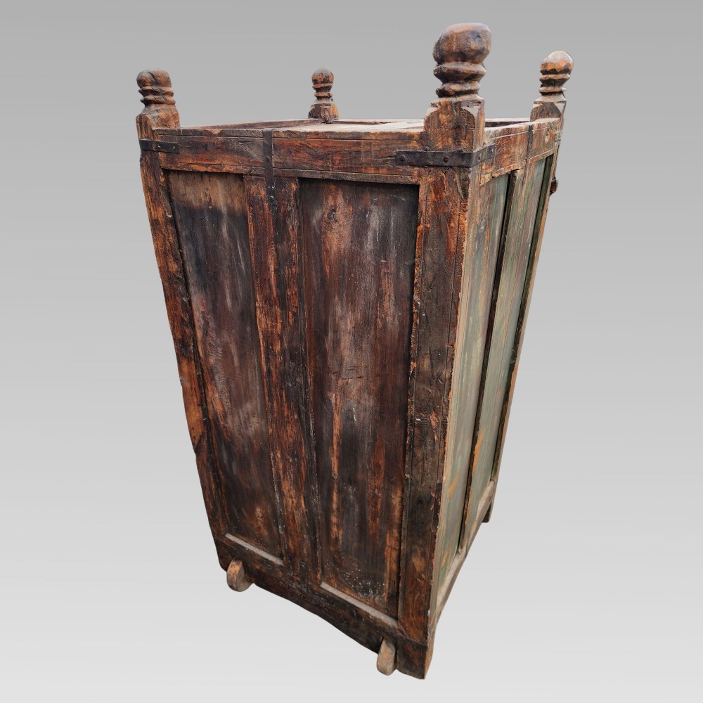 A rustic wooden Indian almirah storage cupboard with distressed green paint, double panel doors, iron fittings, turned corner finials, and wooden wheels. View X