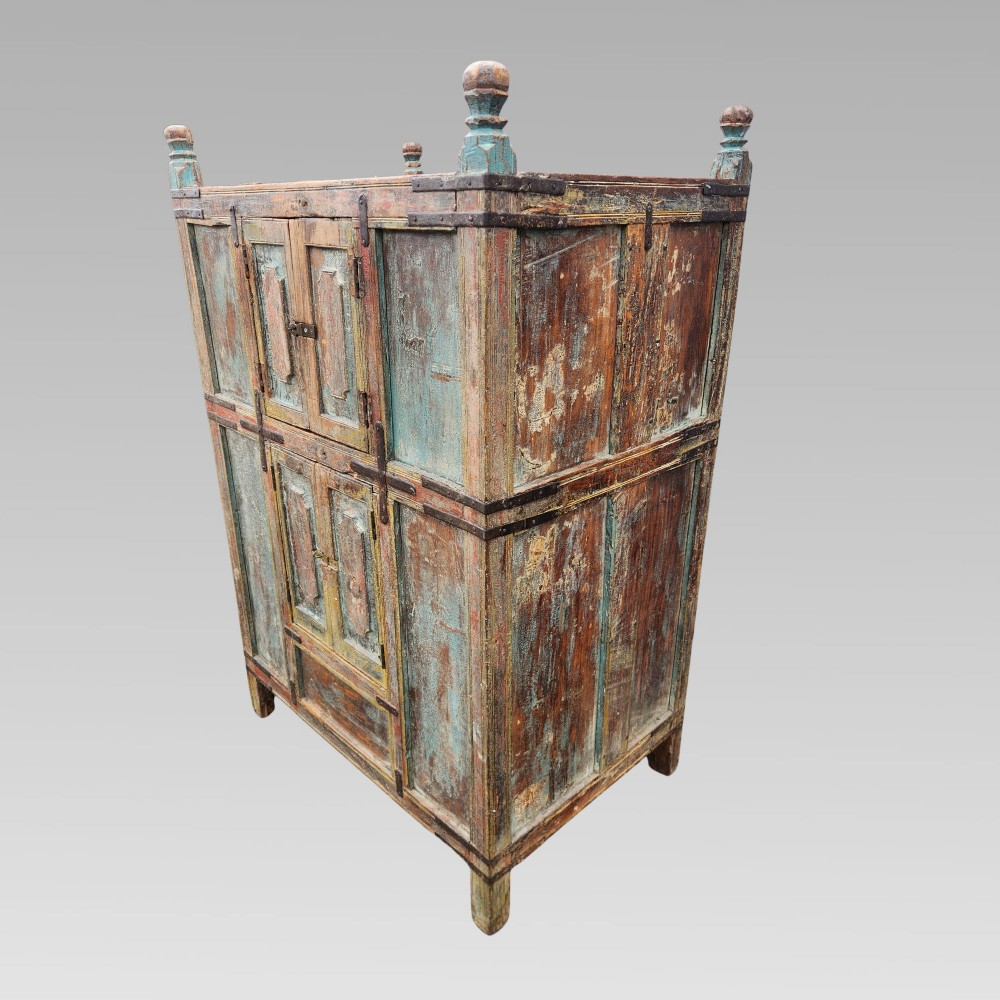 A tall antique Indian wooden almirah cabinet featuring layered distressed blue paint, iron strapping, panelled doors, and corner finials. View V