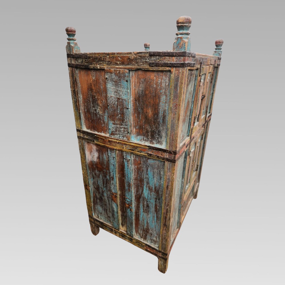 A tall antique Indian wooden almirah cabinet featuring layered distressed blue paint, iron strapping, panelled doors, and corner finials. View X
