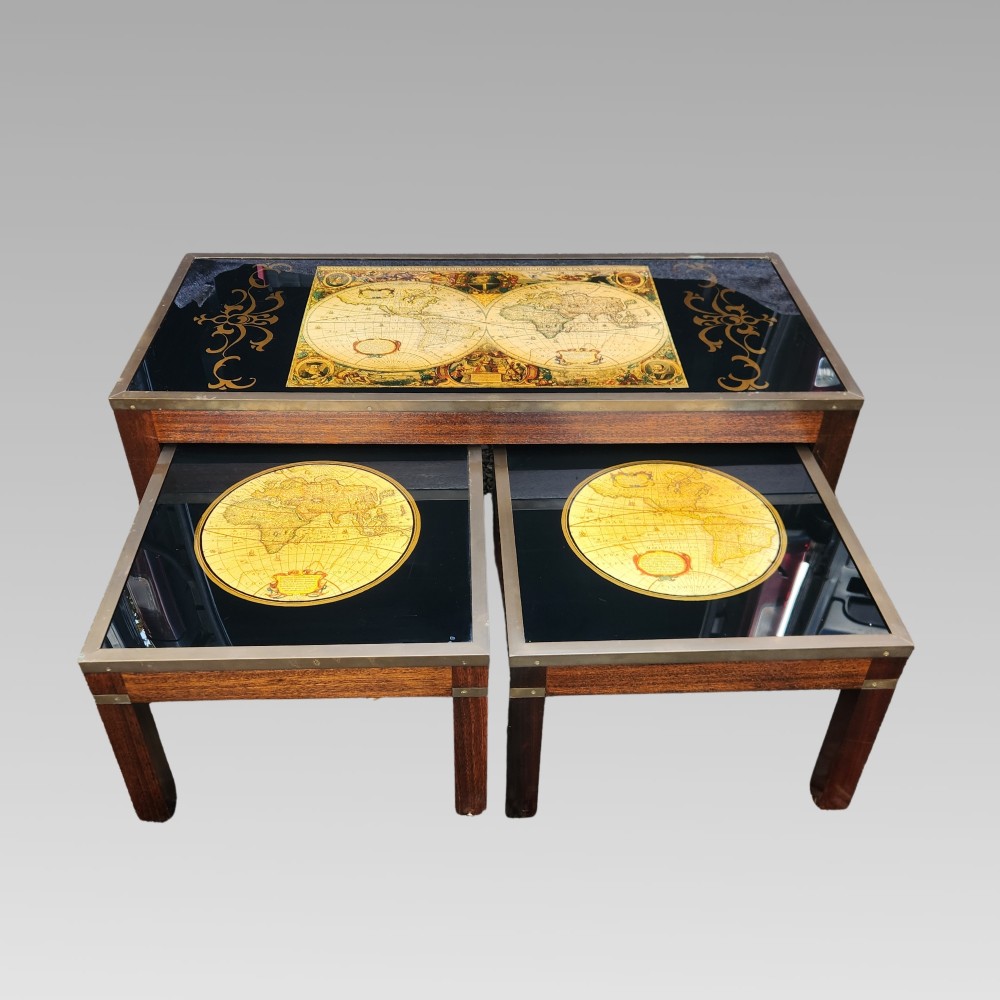 A dark mahogany campaign style nest of three coffee tables featuring glass tops with inset world map illustrations and brass corner mounts.