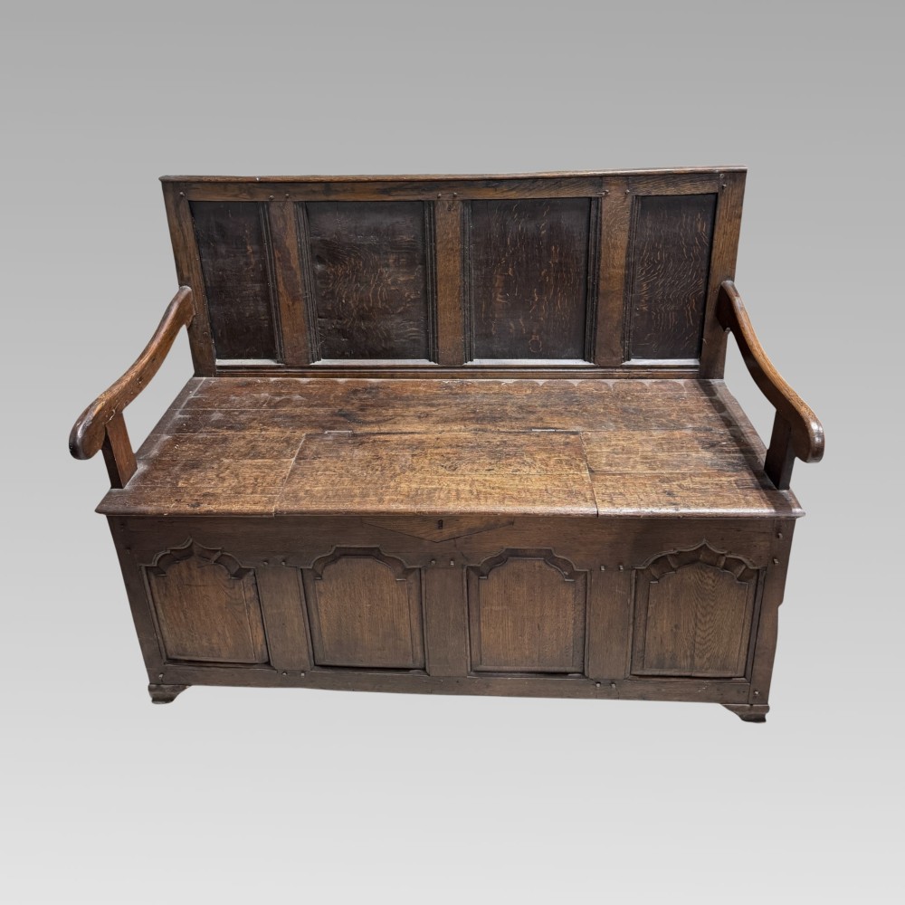 An 18th-century dark oak bench settle with a panelled backrest, shaped open armrests, and an enclosed box base featuring carved arched panels.