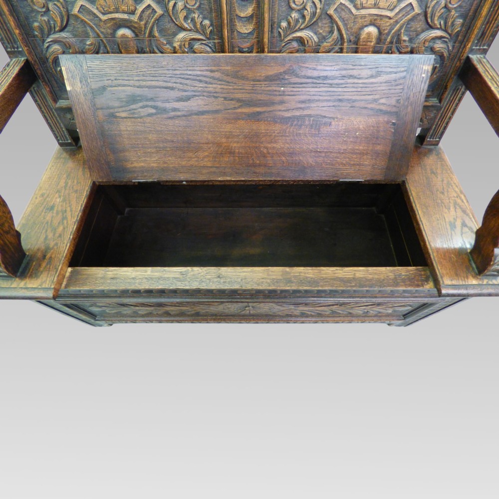 Carved dark oak hall settle featuring a cresting rail, panelled back, curved armrests, and a hinged storage seat. View II