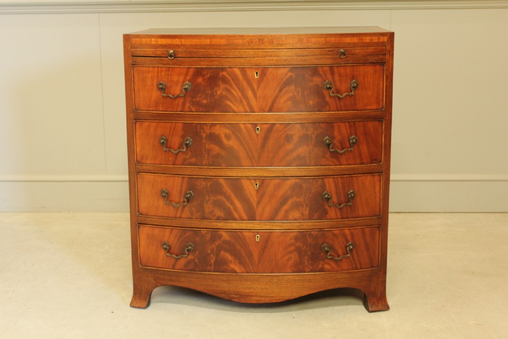 An early 20th-century mahogany bow-fronted bachelors chest of drawers featuring four graduated drawers, a top brushing slide, flame mahogany veneer, and brass handles.