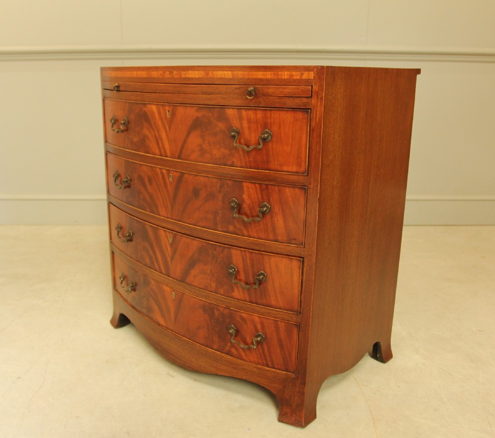 An early 20th-century mahogany bow-fronted bachelors chest of drawers featuring four graduated drawers, a top brushing slide, flame mahogany veneer, and brass handles. View V