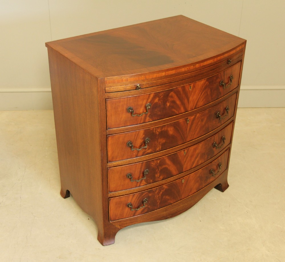 An early 20th-century mahogany bow-fronted bachelors chest of drawers featuring four graduated drawers, a top brushing slide, flame mahogany veneer, and brass handles. View VI