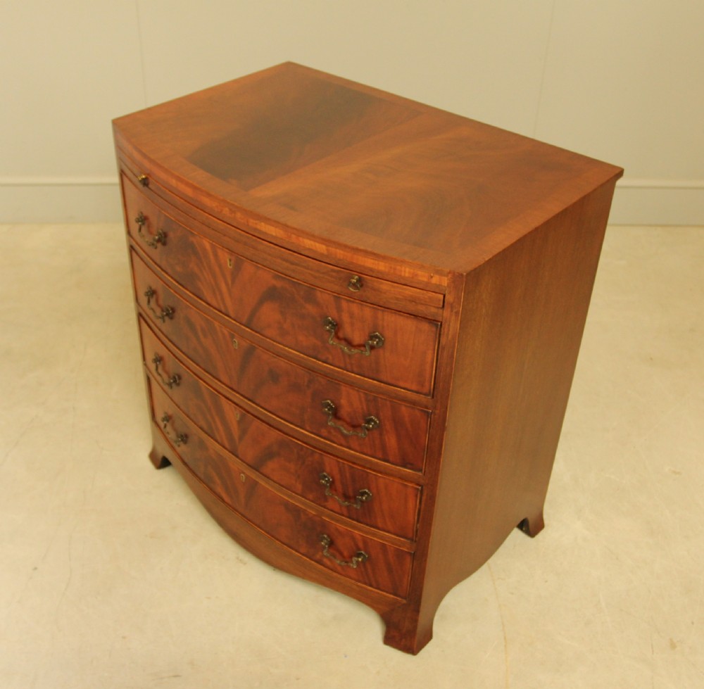 An early 20th-century mahogany bow-fronted bachelors chest of drawers featuring four graduated drawers, a top brushing slide, flame mahogany veneer, and brass handles. View VII