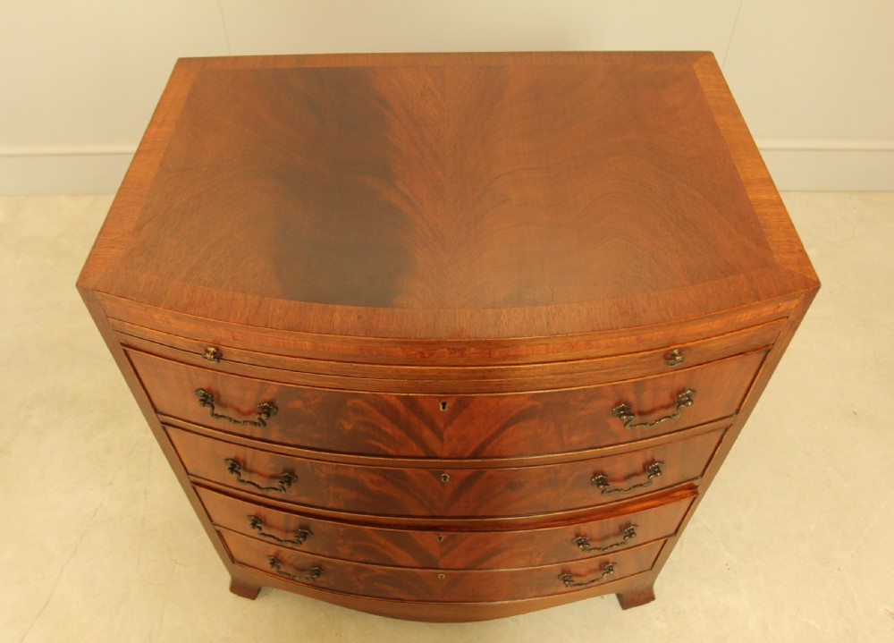 An early 20th-century mahogany bow-fronted bachelors chest of drawers featuring four graduated drawers, a top brushing slide, flame mahogany veneer, and brass handles. View IX