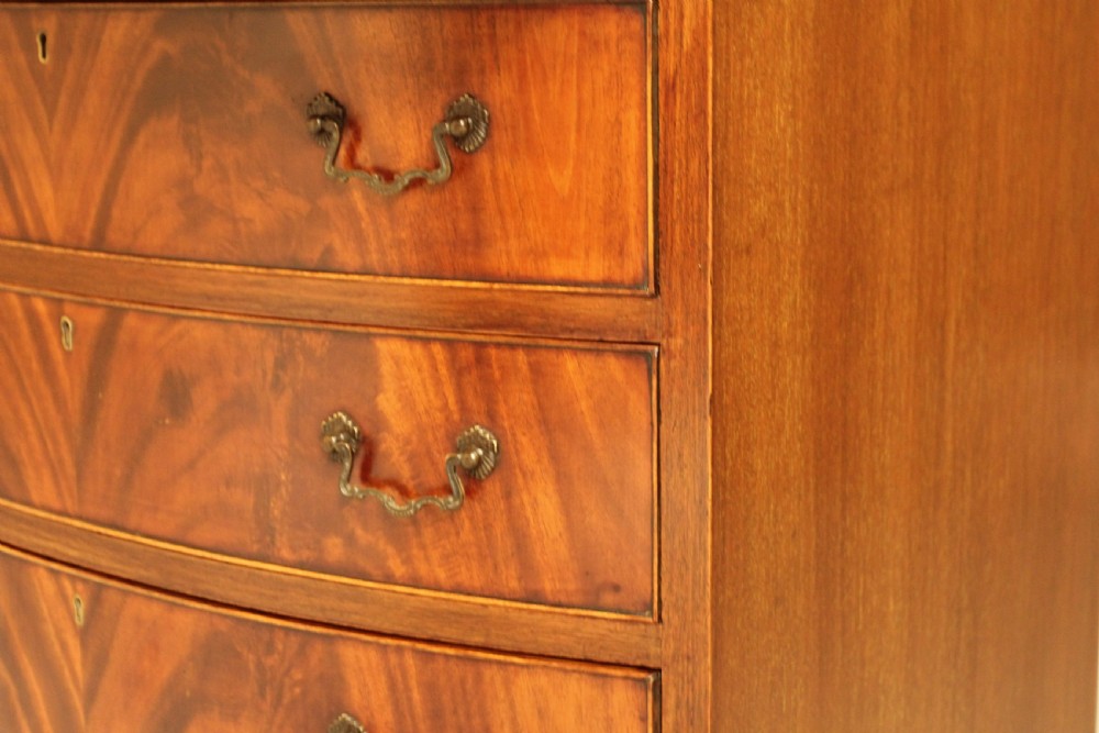 An early 20th-century mahogany bow-fronted bachelors chest of drawers featuring four graduated drawers, a top brushing slide, flame mahogany veneer, and brass handles. View X