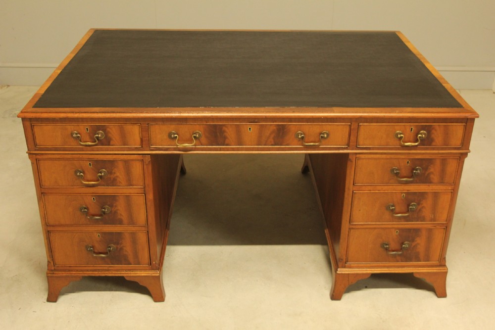 An early twentieth-century curl mahogany partners desk featuring a dark leather writing top, multiple pedestal drawers with brass swan-neck handles, and bracket feet. View III
