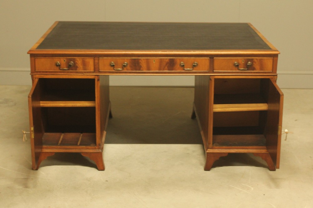 An early twentieth-century curl mahogany partners desk featuring a dark leather writing top, multiple pedestal drawers with brass swan-neck handles, and bracket feet. View IX