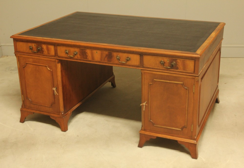 An early twentieth-century curl mahogany partners desk featuring a dark leather writing top, multiple pedestal drawers with brass swan-neck handles, and bracket feet. View X