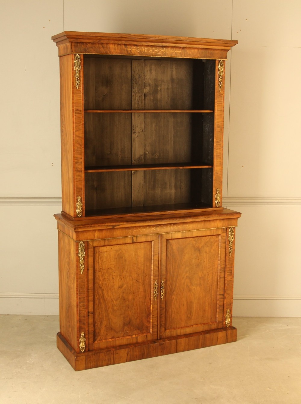 A 19th-century Victorian walnut bookcase featuring an open two-shelf upper section above a two-door lower cupboard with gilt metal mounts and inlaid detailing. View VI