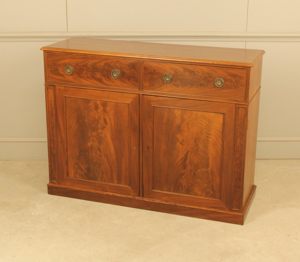 An Edwardian curl mahogany two-door sideboard featuring two top drawers with brass ring handles and panelled cupboard doors on a plinth base. View VI