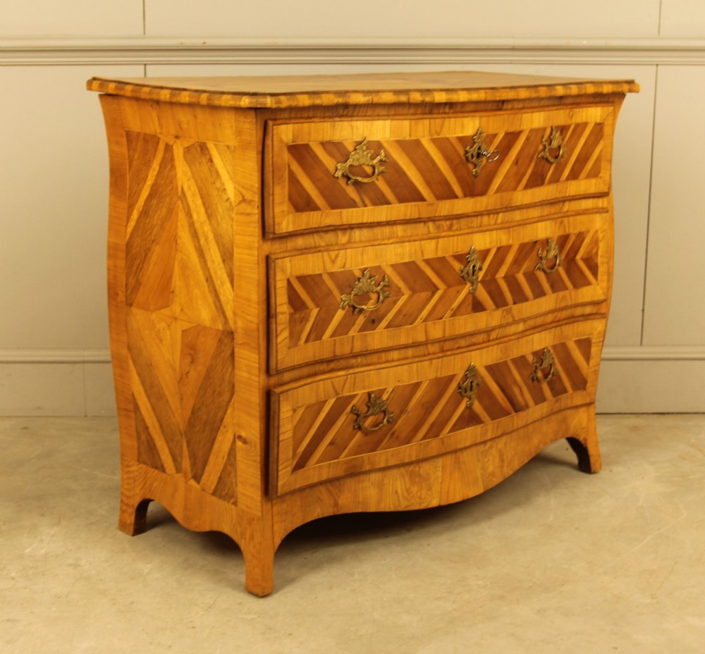 An 18th-century Swedish Rococo bombe chest of drawers featuring geometric chevron parquetry inlays, curved sides, three long drawers, and cast brass hardware. View X