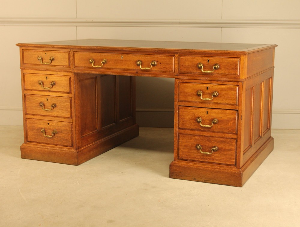 An early 20th-century golden oak twin-pedestal bankers desk featuring a dark leather inset top, nine drawers with brass handles, and panelled sides. View X
