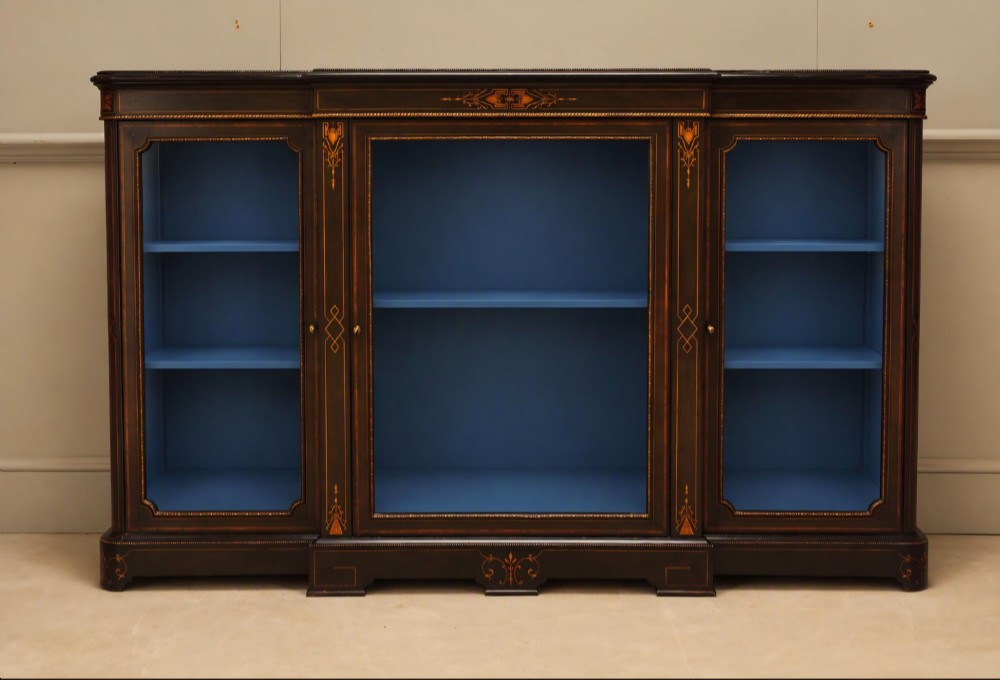 A Victorian ebonised wood breakfront display cabinet featuring three glazed doors, gold inlay detail, and a blue painted interior.