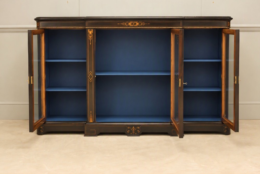 A Victorian ebonised wood breakfront display cabinet featuring three glazed doors, gold inlay detail, and a blue painted interior. View III