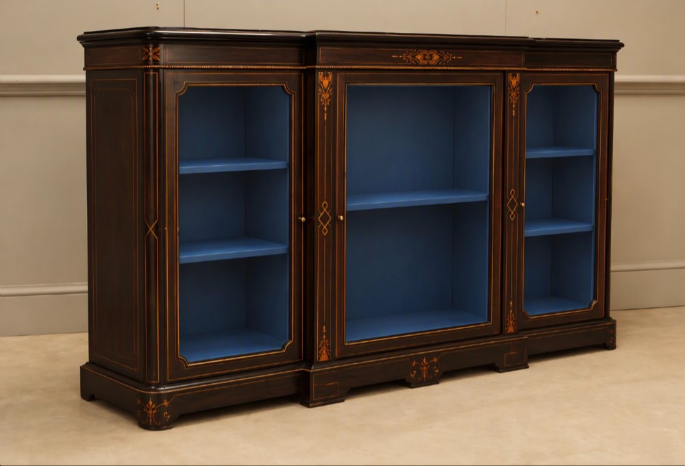 A Victorian ebonised wood breakfront display cabinet featuring three glazed doors, gold inlay detail, and a blue painted interior. View IV