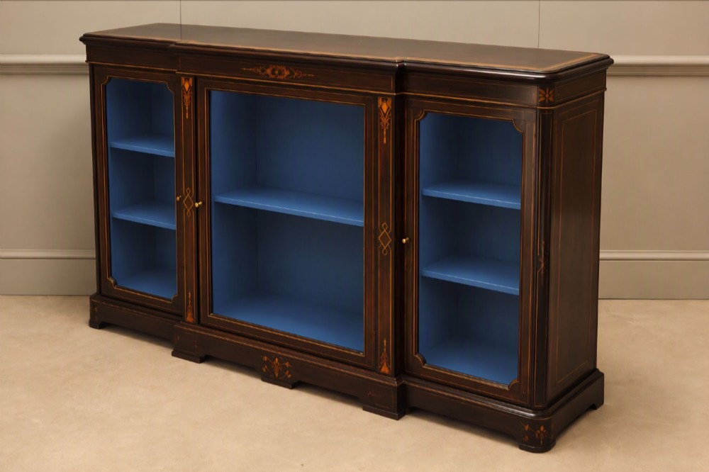 A Victorian ebonised wood breakfront display cabinet featuring three glazed doors, gold inlay detail, and a blue painted interior. View VI