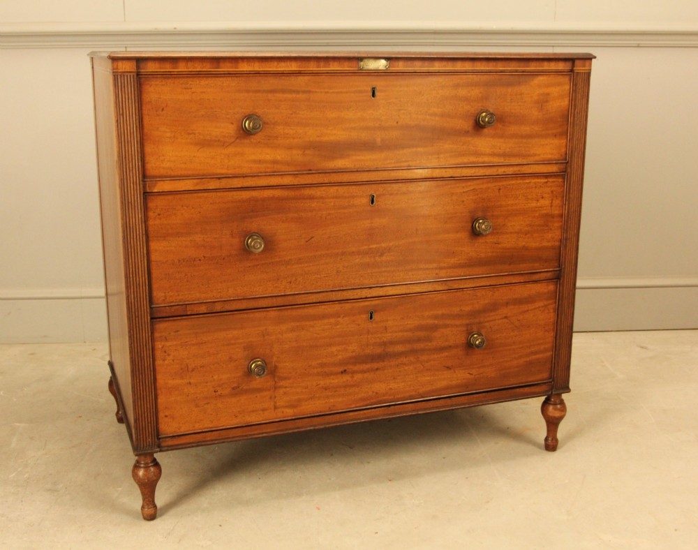 A Regency mahogany chest of drawers featuring three graduated drawers with brass turned knobs and reeded side pilasters resting on turned feet. View II