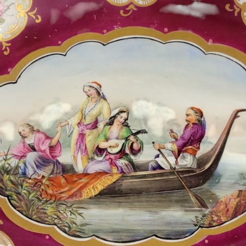 fig 1. An oval early 19th-century claret porcelain serving dish featuring gold scroll handles, floral border panels, and a central hand-painted scene of figures in a boat.