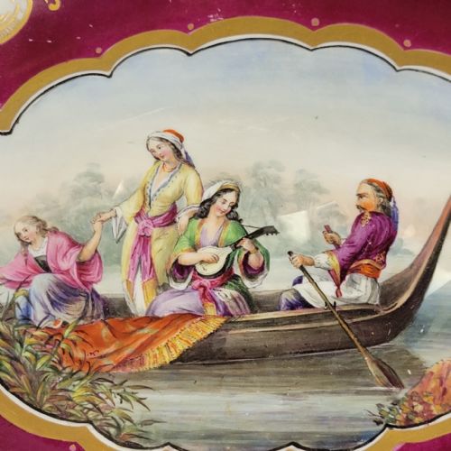 fig 2. An oval early 19th-century claret porcelain serving dish featuring gold scroll handles, floral border panels, and a central hand-painted scene of figures in a boat.
