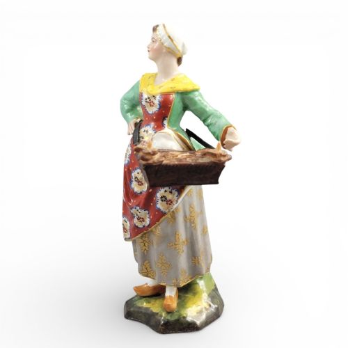 fig 1. An antique Georgian Derby porcelain figurine depicting a female street vendor wearing a green jacket and patterned apron while holding a tray of baked goods.