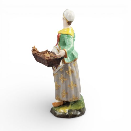 fig 2. An antique Georgian Derby porcelain figurine depicting a female street vendor wearing a green jacket and patterned apron while holding a tray of baked goods.