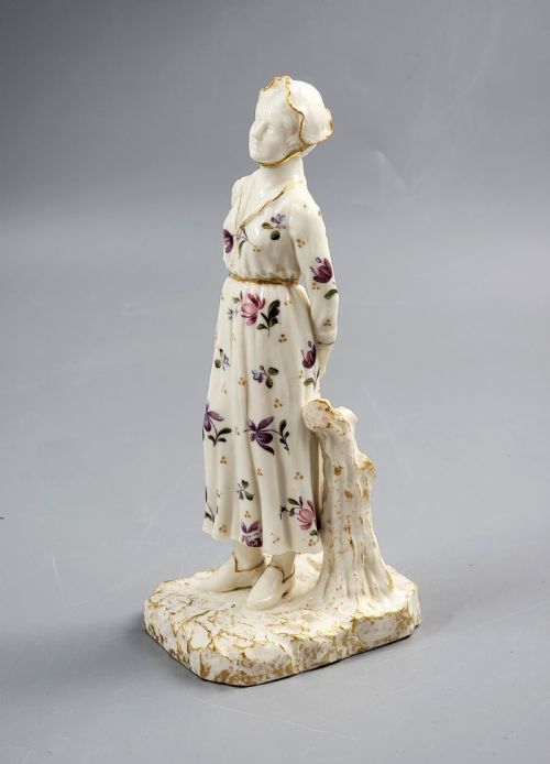 fig 1. A standing porcelain figurine depicting a lady in a floral dress and bonnet with gilded trim on a textured base.