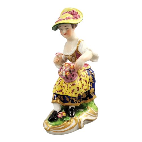 fig 1. An eighteenth-century Derby porcelain figurine depicting a seated female flower seller wearing a yellow bonnet and patterned skirt while holding a small basket of flowers.
