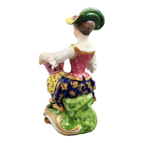 fig 3. An eighteenth-century Derby porcelain figurine depicting a seated female flower seller wearing a yellow bonnet and patterned skirt while holding a small basket of flowers.