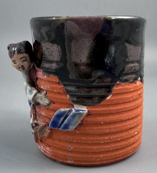 fig 1. A heavy earthenware mug featuring a ribbed terracotta lower section, dark black glaze dripping from the rim, and an applied ceramic figure of a boy in a blue and white robe climbing the side.
