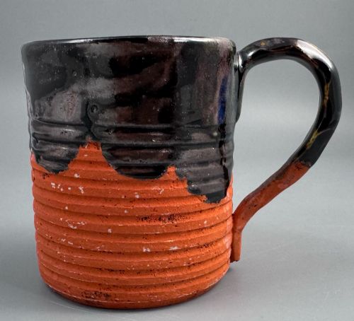 fig 2. A heavy earthenware mug featuring a ribbed terracotta lower section, dark black glaze dripping from the rim, and an applied ceramic figure of a boy in a blue and white robe climbing the side.