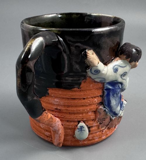 fig 3. A heavy earthenware mug featuring a ribbed terracotta lower section, dark black glaze dripping from the rim, and an applied ceramic figure of a boy in a blue and white robe climbing the side.