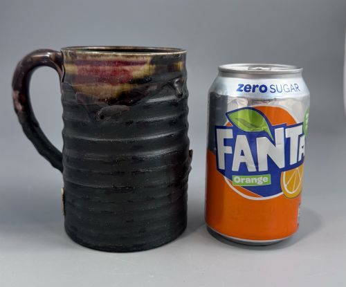 fig 1. A dark ribbed ceramic mug featuring a dripping flambe glaze rim and an applied relief figure of a seated Japanese sennin.