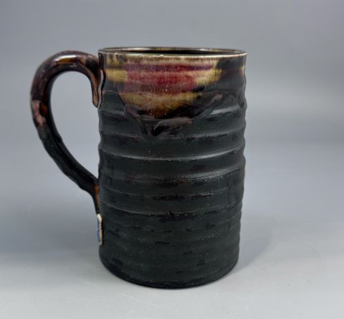 fig 2. A dark ribbed ceramic mug featuring a dripping flambe glaze rim and an applied relief figure of a seated Japanese sennin.