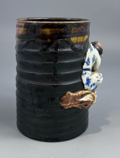fig 3. A dark ribbed ceramic mug featuring a dripping flambe glaze rim and an applied relief figure of a seated Japanese sennin.