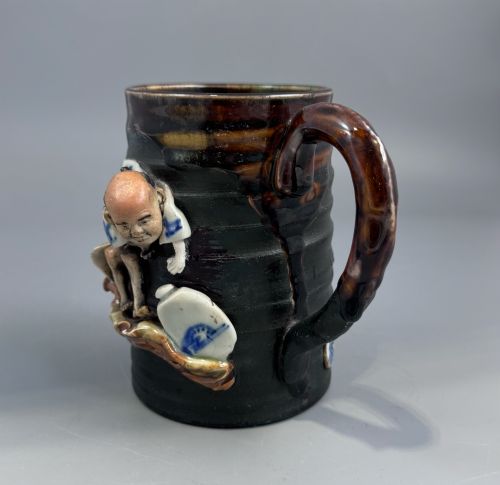 fig 4. A dark ribbed ceramic mug featuring a dripping flambe glaze rim and an applied relief figure of a seated Japanese sennin.