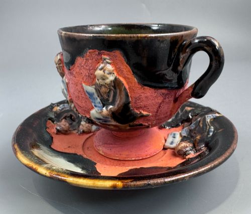 fig 2. A Japanese Sumida Gawa teacup and saucer featuring relief-carved sage figures on a textured red background with a heavy dark flambe glaze.