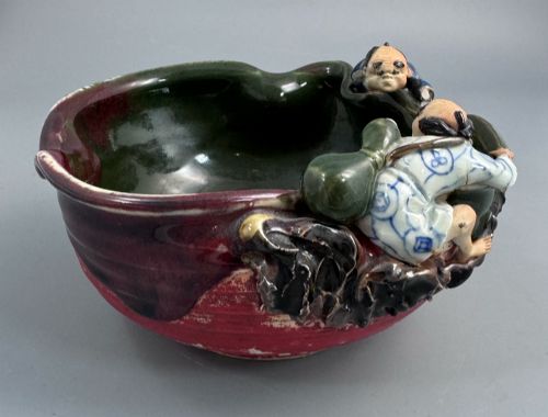 fig 1. A Japanese Sumida Gawa pottery bowl featuring a dark green running glaze over a red base with two sculpted figures leaning over the rim and an applied white makers mark.