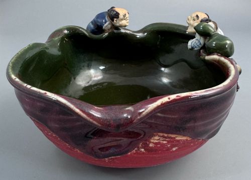 fig 2. A Japanese Sumida Gawa pottery bowl featuring a dark green running glaze over a red base with two sculpted figures leaning over the rim and an applied white makers mark.